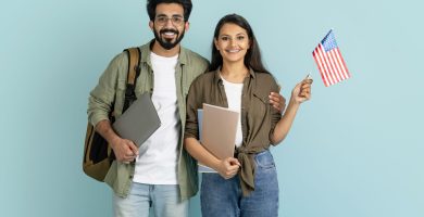 Cheerful happy beautiful multiethnic millennial couple arab man indian woman students with backpack, laptop and notepads showing flag of US, studying abroad, learning English, blue studio background